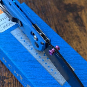 Puede incluir: Una navaja plegable azul con una hoja negra y detalles morados. La navaja est&aacute; sobre una caja azul con el texto "KNIFE COMPANY" y "CPM-S30V". La navaja tiene un clip para el bolsillo.