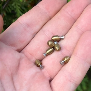 Pode incluir: Um close-up de uma mão segurando seis pequenos parafusos de metal dourados com cabeças prateadas.