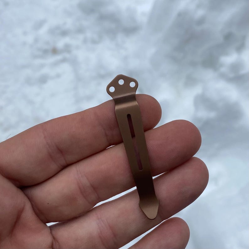 May include: A bronze-colored pocket clip with three small holes at the top and a long, narrow slot down the center. The clip is held in a hand against a snowy background.