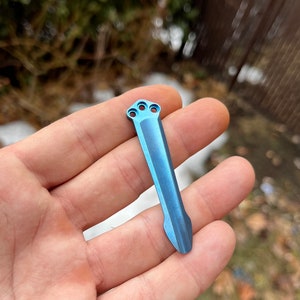 May include: A blue titanium pry bar tool with a pointed tip and three holes at the top.