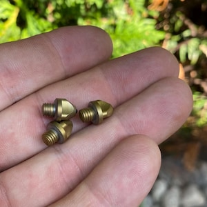 May include: Three brass-colored metal objects with threaded ends and conical heads are displayed in a hand. The objects have a small, circular rubber ring near the base of the head. The background is blurred green foliage.