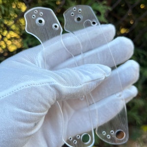 Puede incluir: Dos escamas de cuchillo de plástico transparente con varios agujeros para montaje. Las escamas se sostienen en un paño blanco.