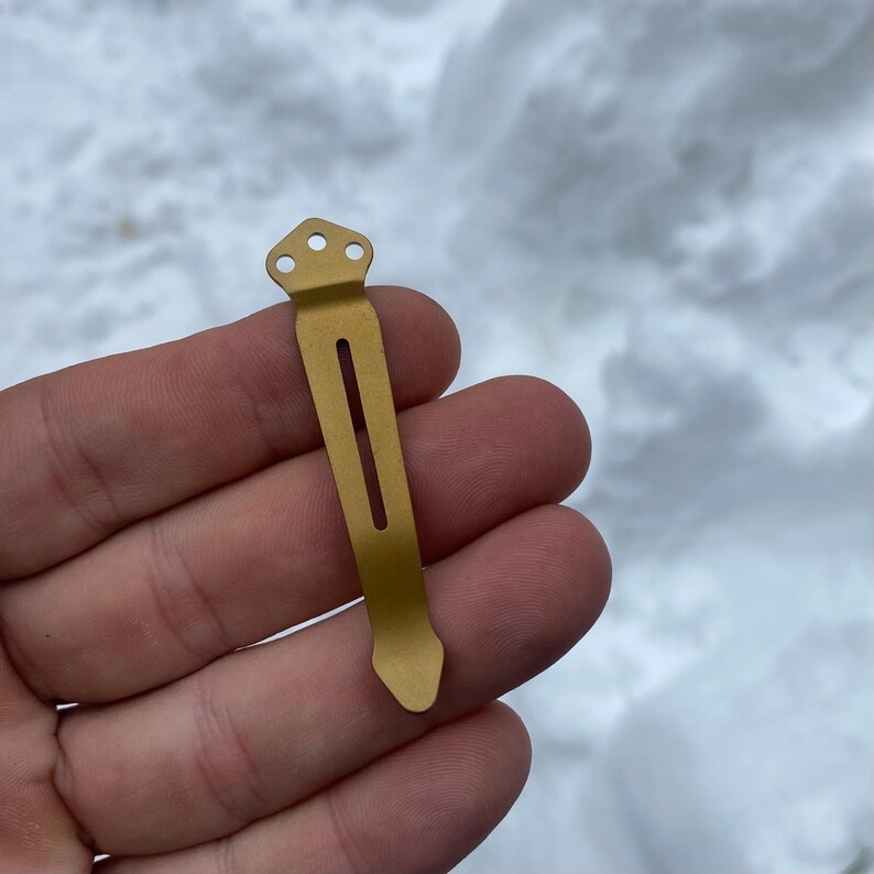 May include: A gold-colored metal pocket clip with three holes at the top and a single slot in the middle.