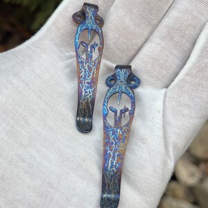 Puede incluir: Dos llaveros de bolsillo de titanio azul y morado con un dise&ntilde;o decorativo. Los llaveros est&aacute;n sobre una superficie blanca.