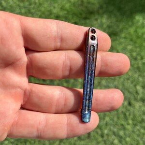 May include: A small, blue and orange metal tool with a textured surface and two holes at the top. The tool is held in a hand.
