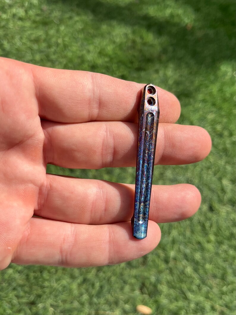 May include: A small, blue and orange metal tool with a textured surface and two holes at the top. The tool is held in a hand.