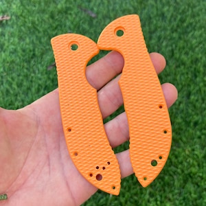 May include: Two orange knife scales with a textured surface and multiple holes. The scales are designed to fit a pocket knife.