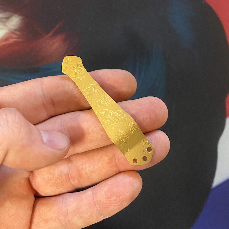 K&ouml;nnte beinhalten: Eine goldfarbene Messerplatine aus Metall mit einem dekorativen Muster und drei L&ouml;chern.