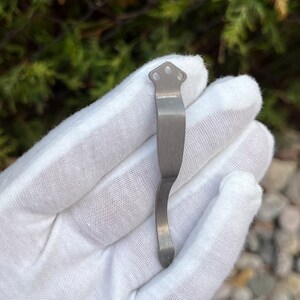 Op de afbeelding: Een zilverkleurige metalen zakclip met een gebogen ontwerp en drie gaten aan de bovenkant.