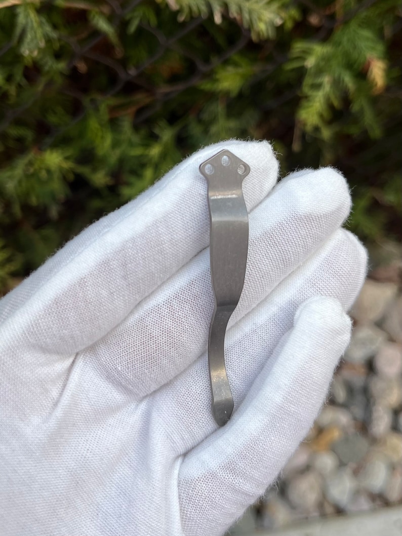 Op de afbeelding: Een zilverkleurige metalen zakclip met een gebogen ontwerp en drie gaten aan de bovenkant.