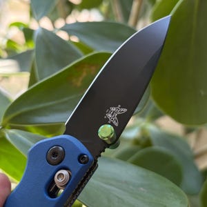 May include: A close-up shot of a black folding knife with a blue handle. The black blade has a silver logo and a green accent. The knife is open, showing the sharp edge.