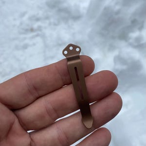 May include: A bronze-colored pocket clip with three small holes at the top and a long, narrow slot down the center. The clip is held in a hand against a snowy background.