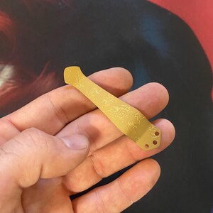 K&ouml;nnte beinhalten: Eine goldfarbene Metall-Messerplatine mit einem filigranen Blumendesign. Die Platine hat drei L&ouml;cher am unteren Ende.