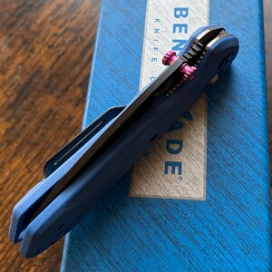 Puede incluir: Una navaja plegable azul con detalles rosas y una hoja plateada. La navaja est&aacute; sobre una caja azul con las palabras "BENCHMADE KNIFE CO." y "MADE" impresas.