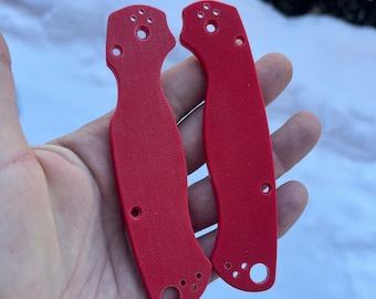 May include: Two bright red knife handle scales with multiple holes for attachment. The scales have a textured surface and are held in a hand against a snowy background. The scales are designed for a folding knife.