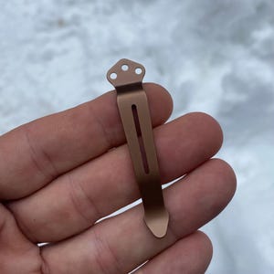 May include: A bronze-colored pocket clip with a rectangular body and a decorative top featuring three small holes. The clip has a streamlined design and is likely for securing a knife or other small tool.