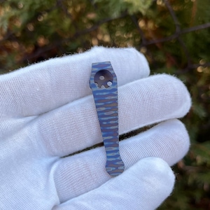 May include: A blue and gray titanium pocket clip with a skull-shaped cutout. The clip is designed to attach to a pocket or belt.
