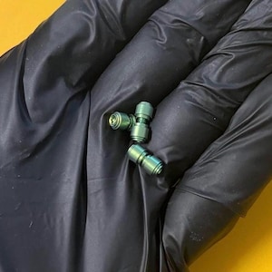 May include: Close-up of three small, metallic green objects. Each object has a cylindrical shape with a ridged design and a slight blue hue. The objects are resting on a black surface with a yellow background.