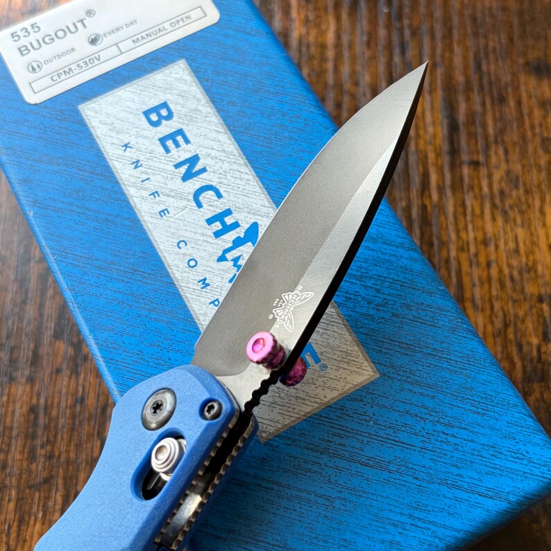 Puede incluir: Primer plano de un cuchillo Benchmade Bugout azul con una hoja plateada. La hoja tiene un logotipo de mariposa y detalles en rosa. El cuchillo est&aacute; parcialmente abierto, mostrando el filo. La caja es azul con texto blanco.