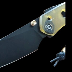 Könnte beinhalten: Ein schwarzes Klappmesser mit einem gelben und goldenen Griff. Der Griff hat ein kreisförmiges Logo mit dem Buchstaben "C" darin.