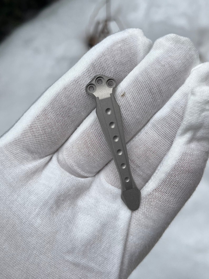 May include: A silver titanium pocket knife handle with a textured surface and a rounded tip.