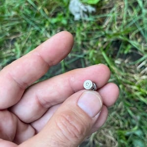 May include: A small, silver-colored metal object with a circular design on its top. The object is held between fingers against a backdrop of green grass.