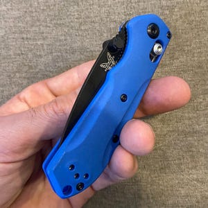 May include: A blue and black folding knife with a black blade and a blue handle. The knife has a butterfly logo and several black screws. The knife is held in a hand.