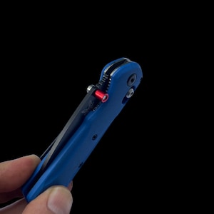 Puede incluir: Un cuchillo de bolsillo azul con una hoja negra y un acento rojo. El cuchillo está parcialmente abierto y la hoja es visible.