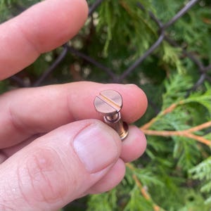 May include: A close-up of a copper-colored screw with a slotted head. The screw is held in a person's hand.