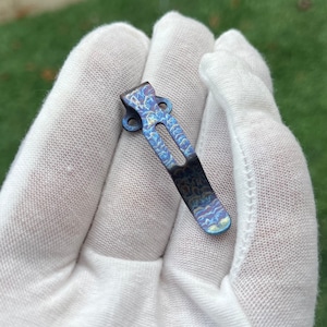 May include: A blue titanium pocket clip with a swirling pattern. The clip is designed to attach to a pocket or belt.