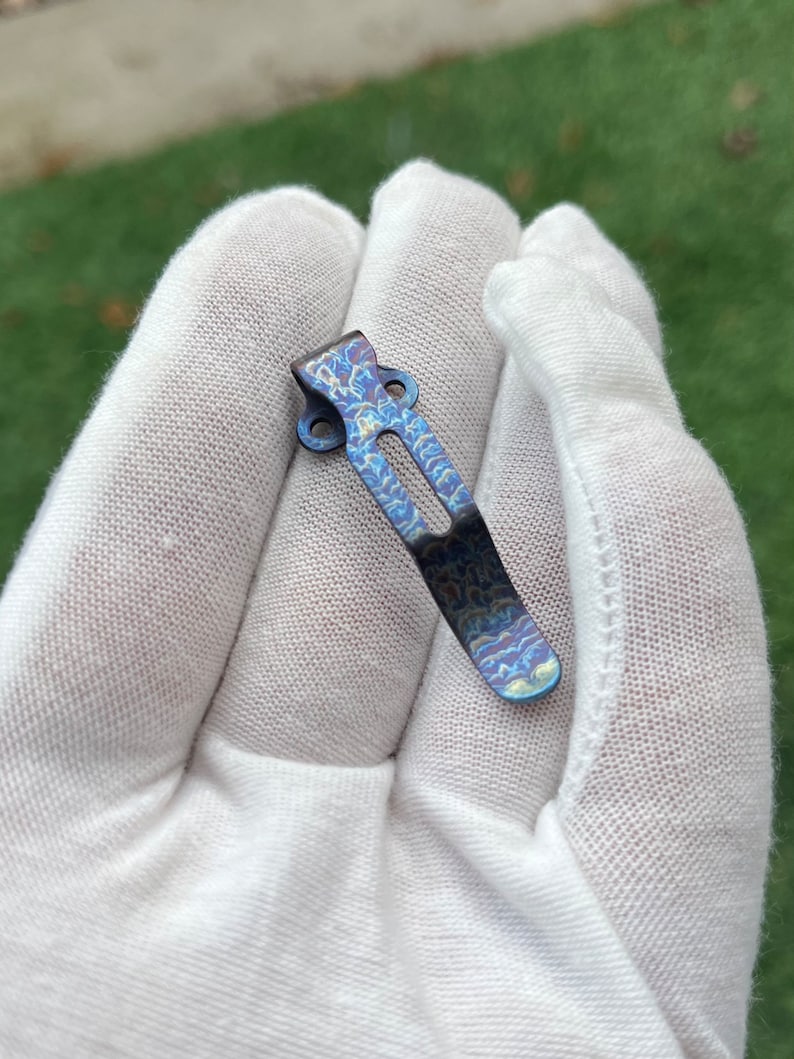 May include: A blue titanium pocket clip with a swirling pattern. The clip is designed to attach to a pocket or belt.