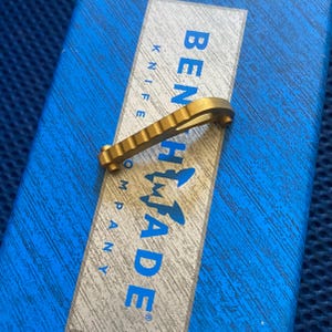 May include: A gold-colored metal knife part with a serrated edge and a curved shape. The part is on a blue box with the text "BENCHMADE KNIFE COMPANY" printed on it.