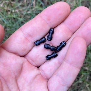 May include: A close-up of several small, black plastic connectors. The connectors are cylindrical with a slightly textured surface and appear to have a series of ridges and grooves. They are held in an open hand against a background of green grass.