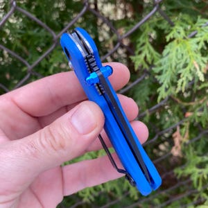 Puede incluir: Un cuchillo de bolsillo azul con una hoja negra y un mango azul. El cuchillo está cerrado y tiene un pequeño botón azul en el lateral.