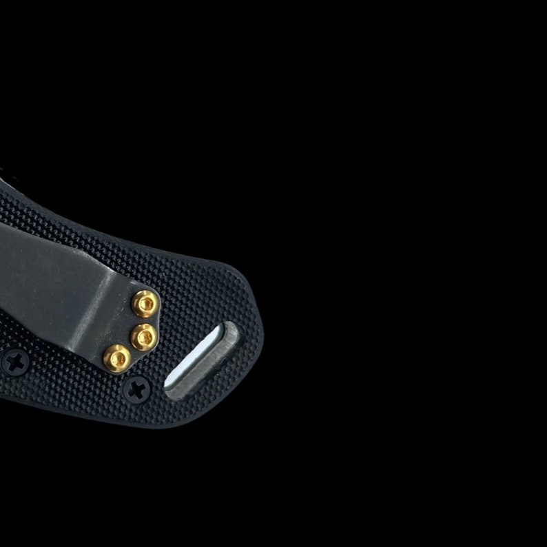 Puede incluir: Primer plano de una navaja de bolsillo negra con mango texturizado y clip. La navaja tiene tornillos de color dorado.