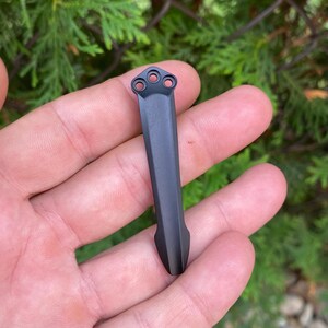May include: A matte black metal tool with a long, rectangular body and a pointed end. The top of the tool features three circular holes. The tool is held in a person's hand against a backdrop of green foliage.
