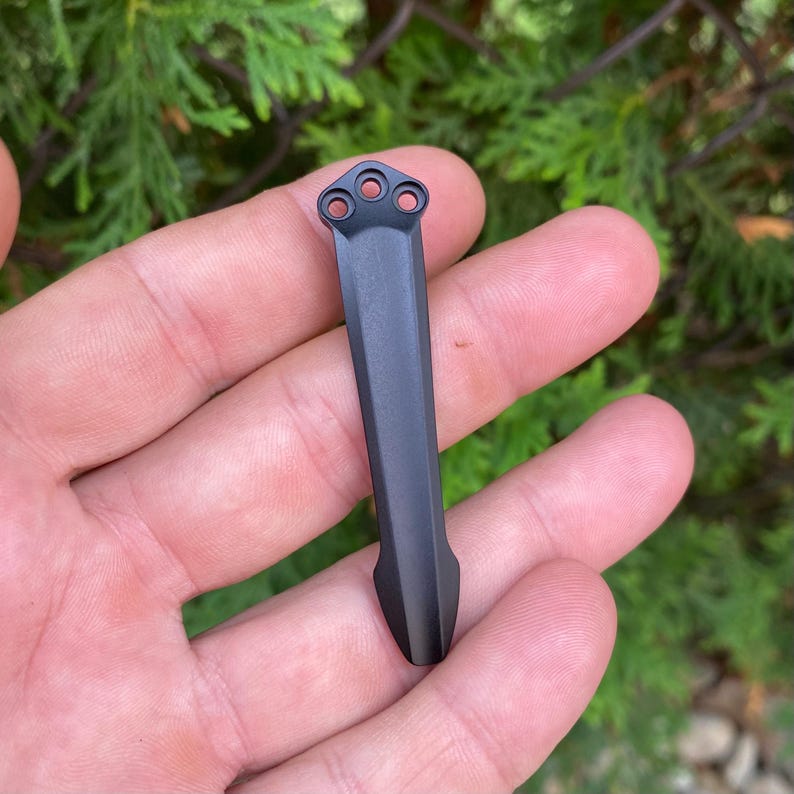 May include: A matte black metal tool with a long, rectangular body and a pointed end. The top of the tool features three circular holes. The tool is held in a person's hand against a backdrop of green foliage.