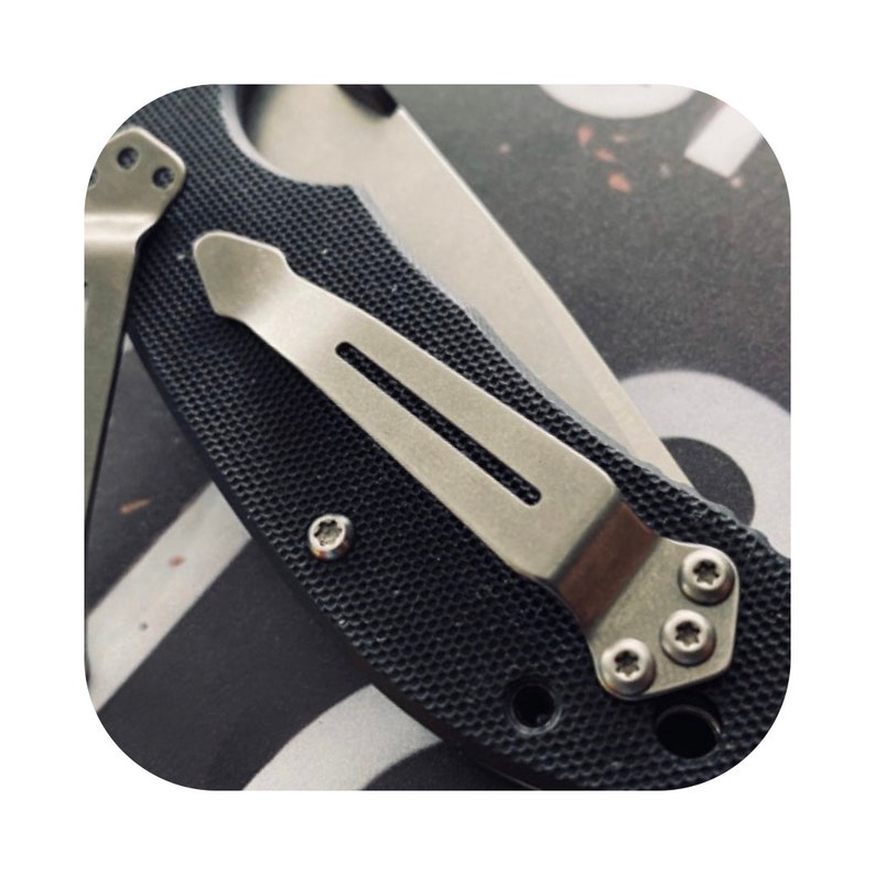 May include: Close-up of a black pocket knife with a silver pocket clip. The clip has a textured surface and is secured with four screws.