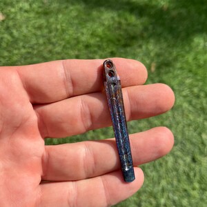 May include: A small, metallic, iridescent blue and copper colored,  knife blade with a textured surface and two holes at the top.
