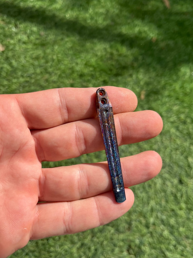 May include: A small, metallic, iridescent blue and copper colored,  knife blade with a textured surface and two holes at the top.