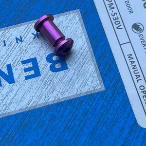 Peut inclure: Une vis métallique violette avec un corps cylindrique et deux extrémités évasées. La vis est positionnée en diagonale sur une surface bleue avec le mot "BEN" en lettres bleues. Une étiquette blanche avec le texte "OUTDOOR" et "CPM-S30V" est visible.