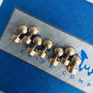 Puede incluir: Seis pomos de metal de color dorado en forma de bola con una base roscada. Los pomos están dispuestos en una fila sobre una superficie azul y blanca con el texto "KNIFE COMPANY" visible.