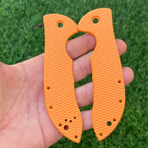 May include: A pair of orange knife scales with a textured surface and multiple holes. The scales are designed to fit a knife handle.