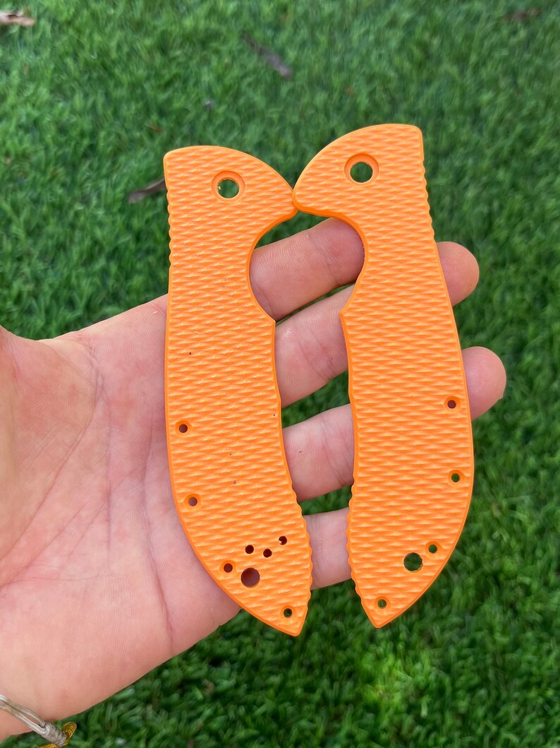 May include: A pair of orange knife scales with a textured surface and multiple holes. The scales are designed to fit a knife handle.