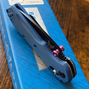 Puede incluir: Una navaja plegable azul con una hoja negra y detalles rosas. La navaja tiene un logotipo de mariposa y est&aacute; parcialmente abierta. Descansa sobre una caja azul. La hoja es visible.