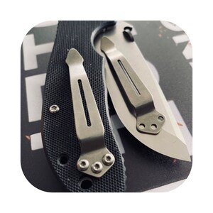May include: Two silver pocket clip attachments for a knife. The clips are attached to a black knife handle with a textured surface.