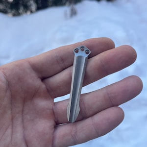 Könnte beinhalten: Ein silbernes Metallwerkzeug mit einer spitzen Spitze und drei Löchern oben. Das Werkzeug wird in einer Hand vor einem schneebedeckten Hintergrund gehalten.