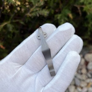 May include: A silver metal pocket clip with two small holes. The clip is curved and has a smooth finish.