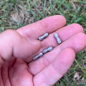 May include: A collection of small, silver-colored metal objects, possibly fasteners or connectors, are held in a person's hand. The objects have a cylindrical shape with a slightly textured surface and appear to be made of metal.