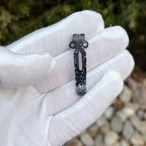 May include: Black metal pocket clip with white speckles. The clip has two holes at the top and a slot in the middle.
